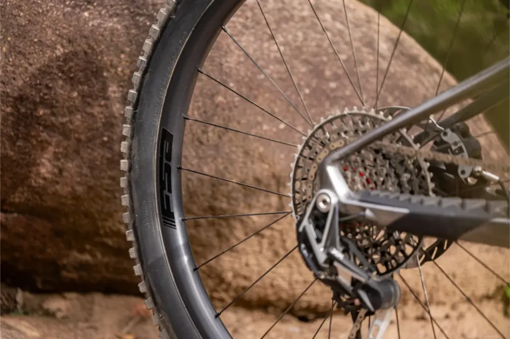 Primer plano de la rueda trasera de una bicicleta de montaña, el cassette y el desviador, con una gran roca al fondo.