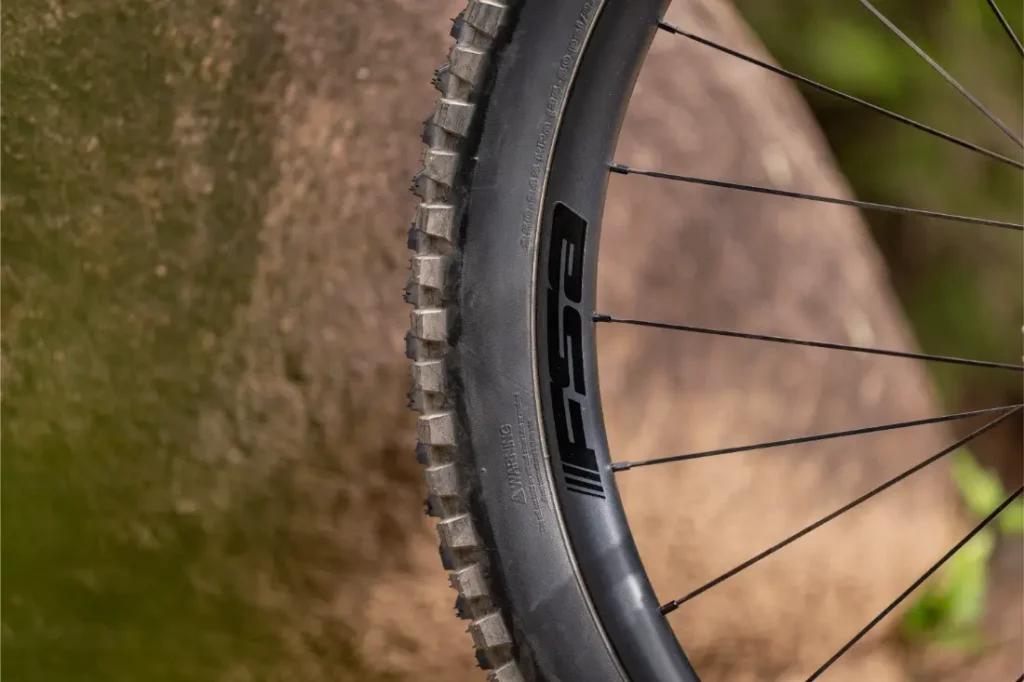 Primer plano de una rueda de bicicleta de montaña y su llanta frente a una gran roca, en el que se aprecia el dibujo de la banda de rodadura y parte de la llanta negra con un logotipo visible.
