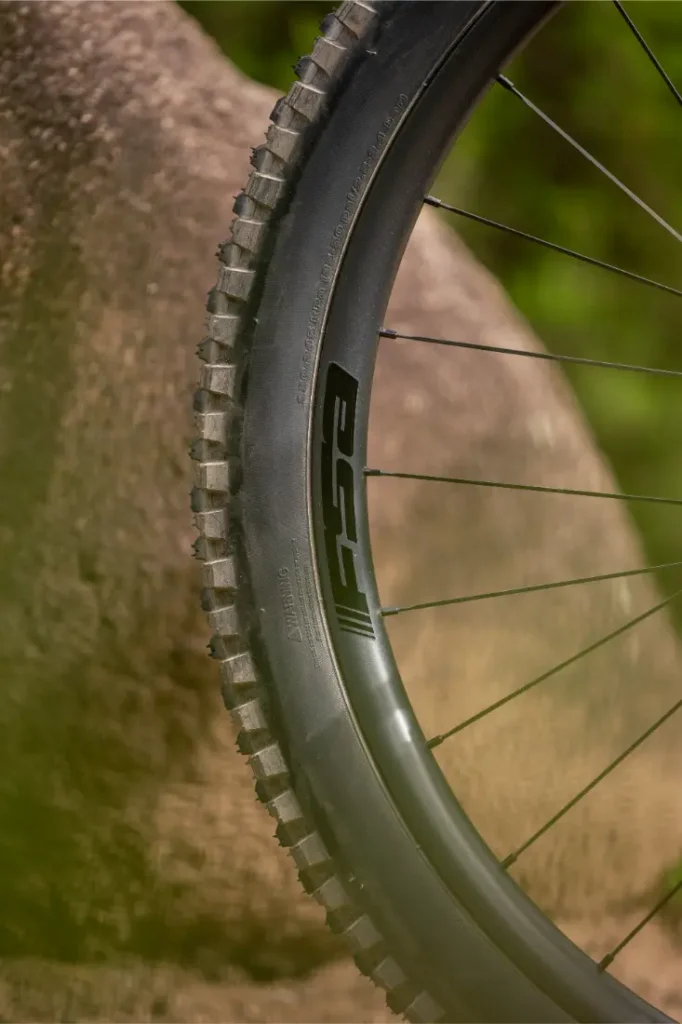 Primer plano de una rueda y una llanta de bicicleta de montaña en las que se aprecia el logotipo «ESC», situadas junto a una gran roca en un entorno al aire libre.