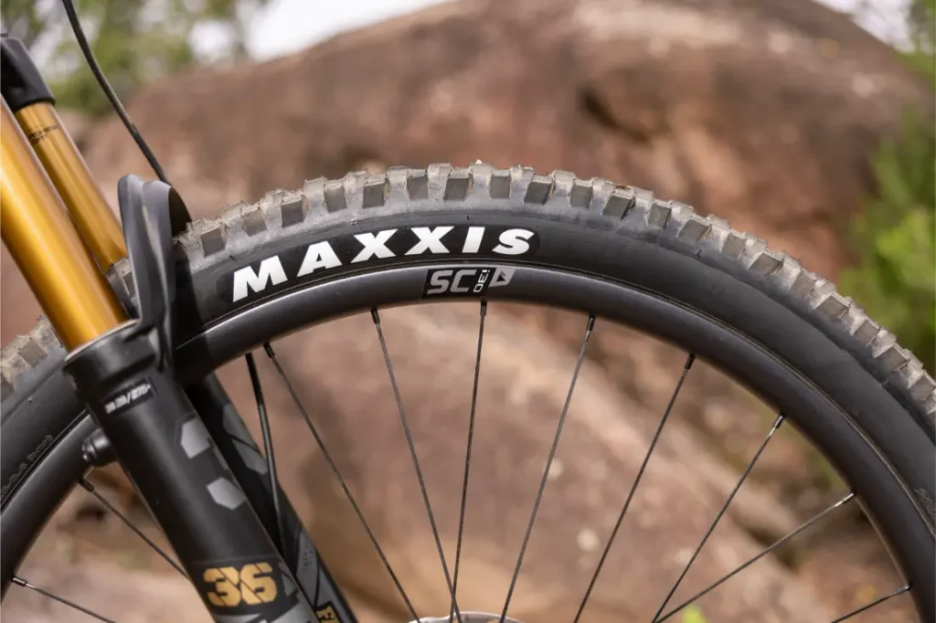 Primer plano de la rueda delantera de una bicicleta de montaña con un neumático Maxxis, radios negros y una horquilla de suspensión dorada, situada frente a una gran roca al aire libre.