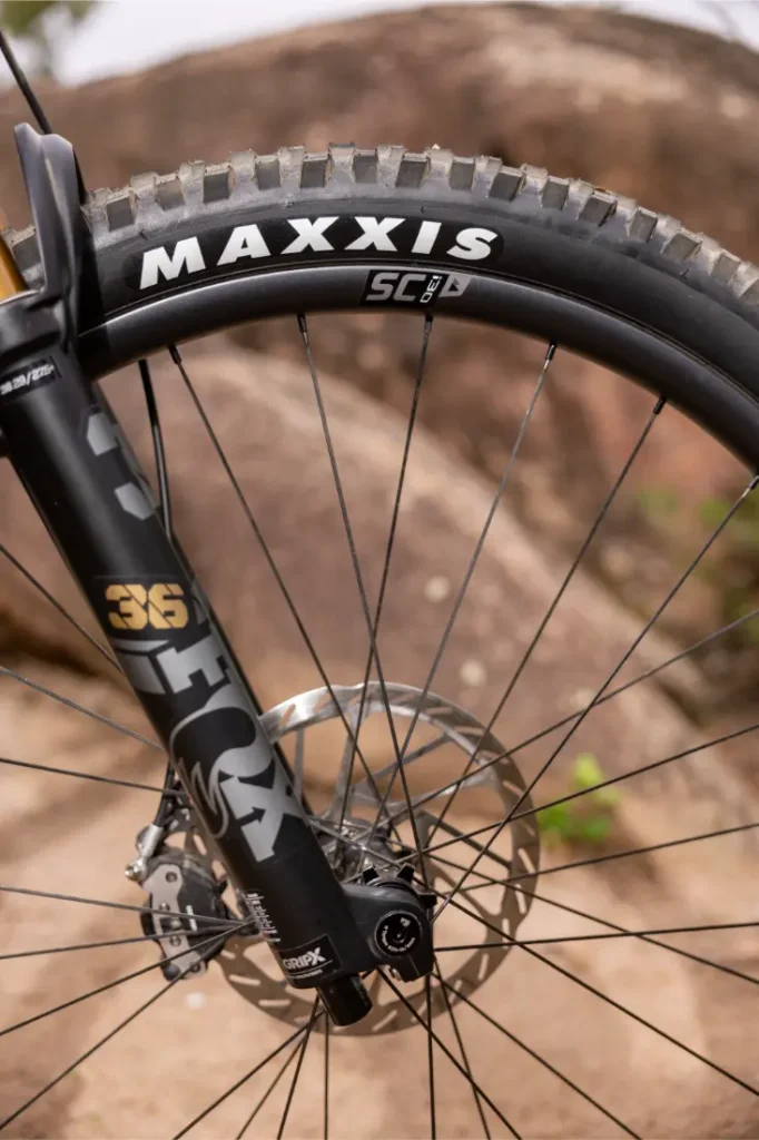 Primer plano de la rueda delantera de una bicicleta de montaña con un neumático Maxxis, un freno de disco y una horquilla de suspensión FOX 36, con un fondo rocoso al aire libre.