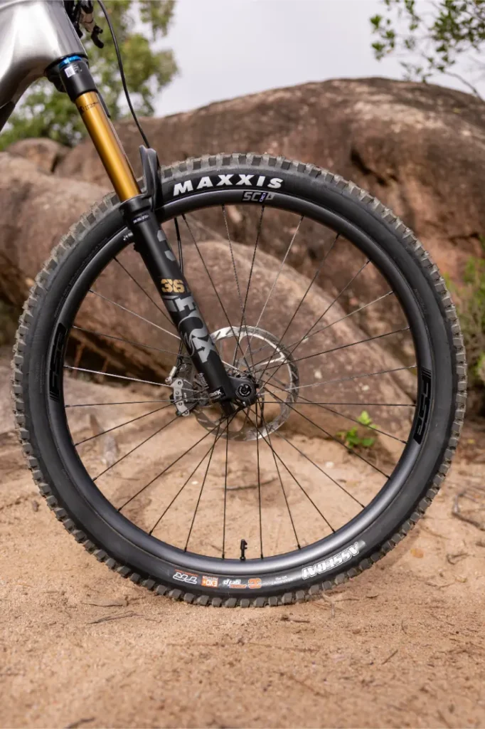 Un primer plano de la rueda delantera de una bicicleta de montaña con un neumático Maxxis, un freno de disco y una horquilla de suspensión, situada en un sendero de tierra con rocas y vegetación al fondo.