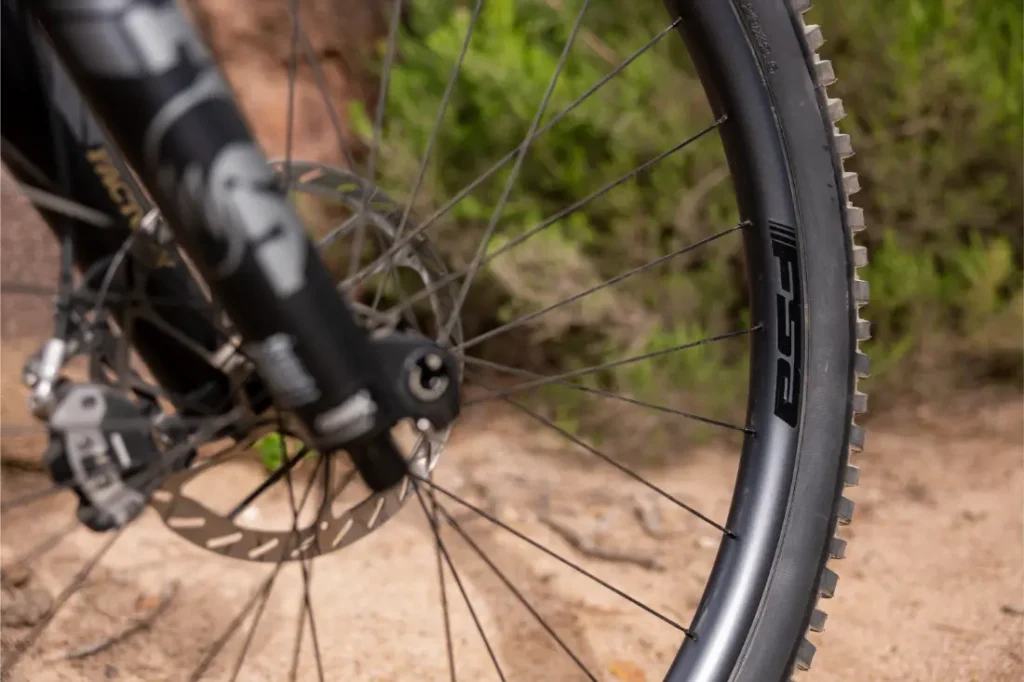 Primer plano de la rueda delantera de una bicicleta con freno de disco y el logotipo de FSA, aparcada en un camino de tierra con vegetación al fondo.