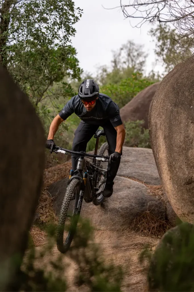 Un ciclista de montaña, con casco y gafas de sol, pedalea sobre las rocas por un sendero de tierra rodeado de grandes cantos rodados y vegetación.