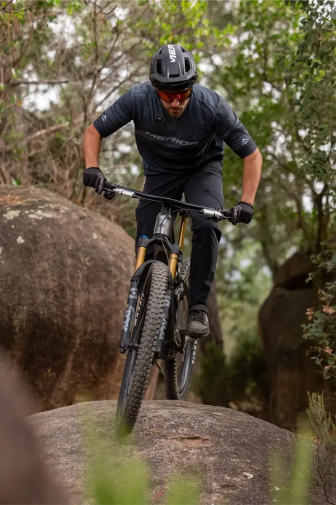 Una persona que lleva casco y gafas de sol va en bicicleta de montaña por encima de una gran roca, rodeada de árboles y cantos rodados.
