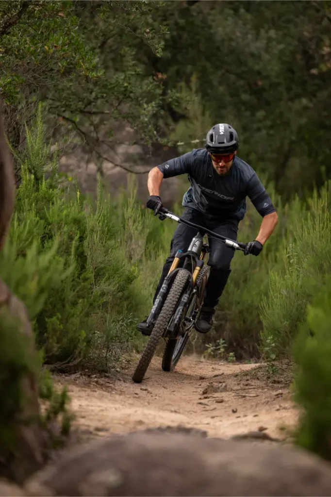 Una persona que lleva casco y gafas de sol baja en bicicleta de montaña por un sendero de tierra rodeado de arbustos verdes y árboles.