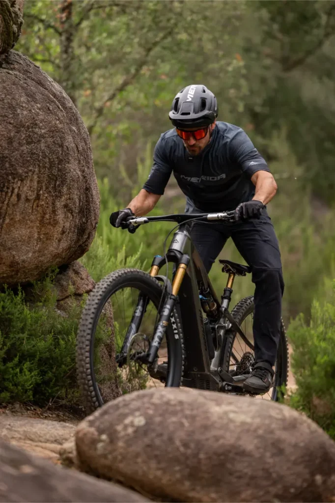 Una persona que lleva casco y gafas de sol va en bicicleta de montaña por un sendero rocoso rodeado de vegetación y grandes rocas.