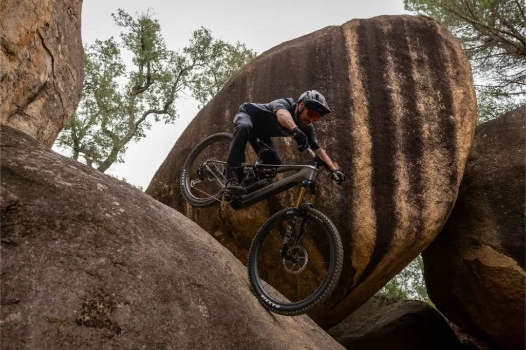 Una persona que lleva casco baja en bicicleta de montaña por una pendiente empinada y rocosa, con grandes rocas y árboles al fondo.
