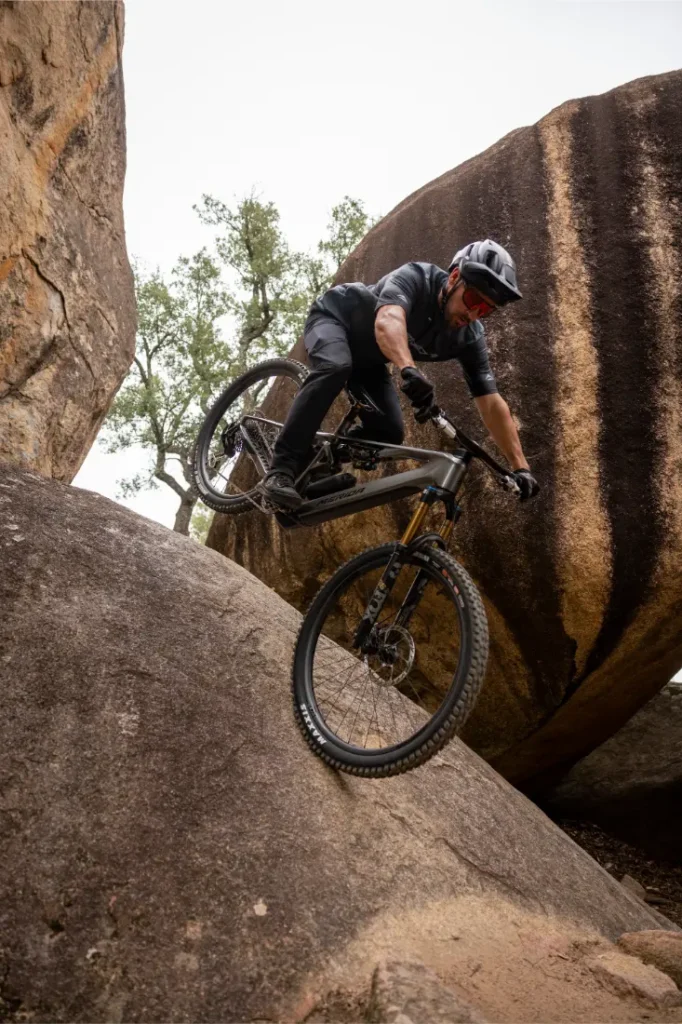 Un ciclista de montaña, con todo el equipo puesto, desciende por una empinada pared rocosa entre grandes cantos rodados, haciendo gala de su control y equilibrio.