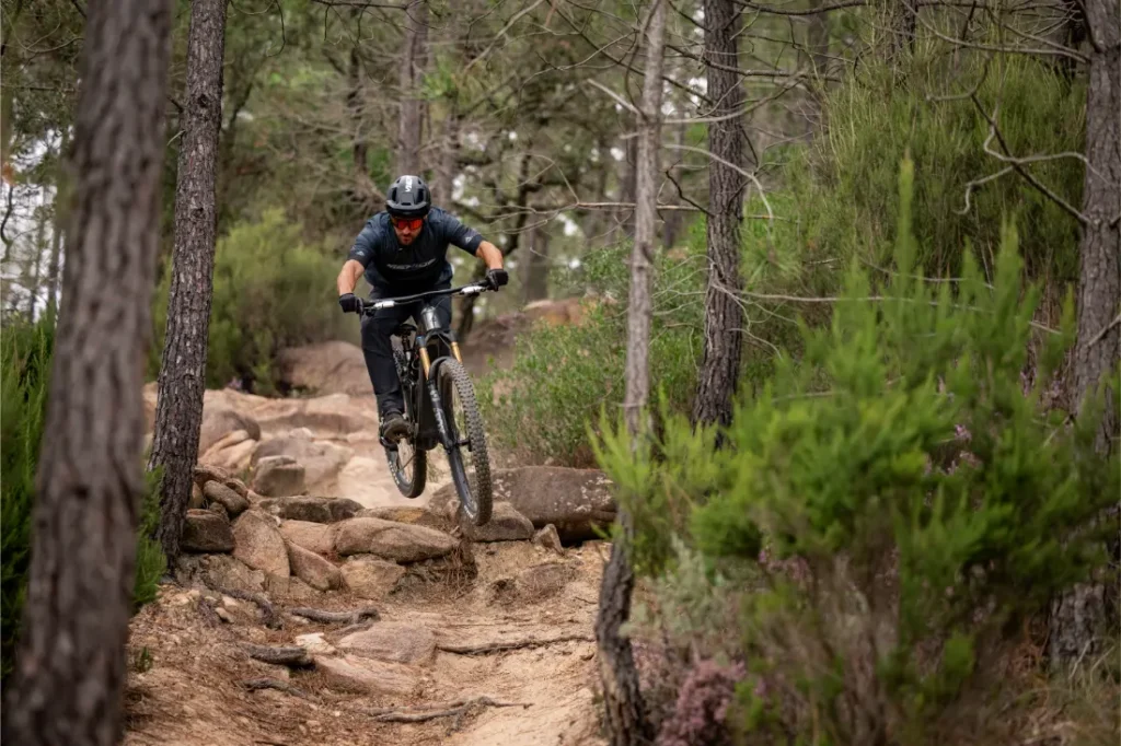 Una persona que lleva casco baja en bicicleta de montaña por un sendero rocoso que atraviesa una zona boscosa con pinos y matorrales.