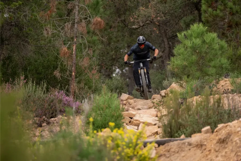 Una persona que lleva casco y equipación de ciclismo recorre en bicicleta de montaña un sendero rocoso rodeado de árboles verdes y flores silvestres.