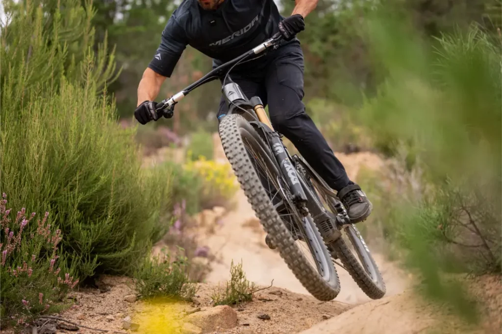 Una persona vestida de negro va en bicicleta de montaña por un camino fuera de pista, levantando la rueda delantera sobre un sendero de tierra rodeado de arbustos y plantas verdes.