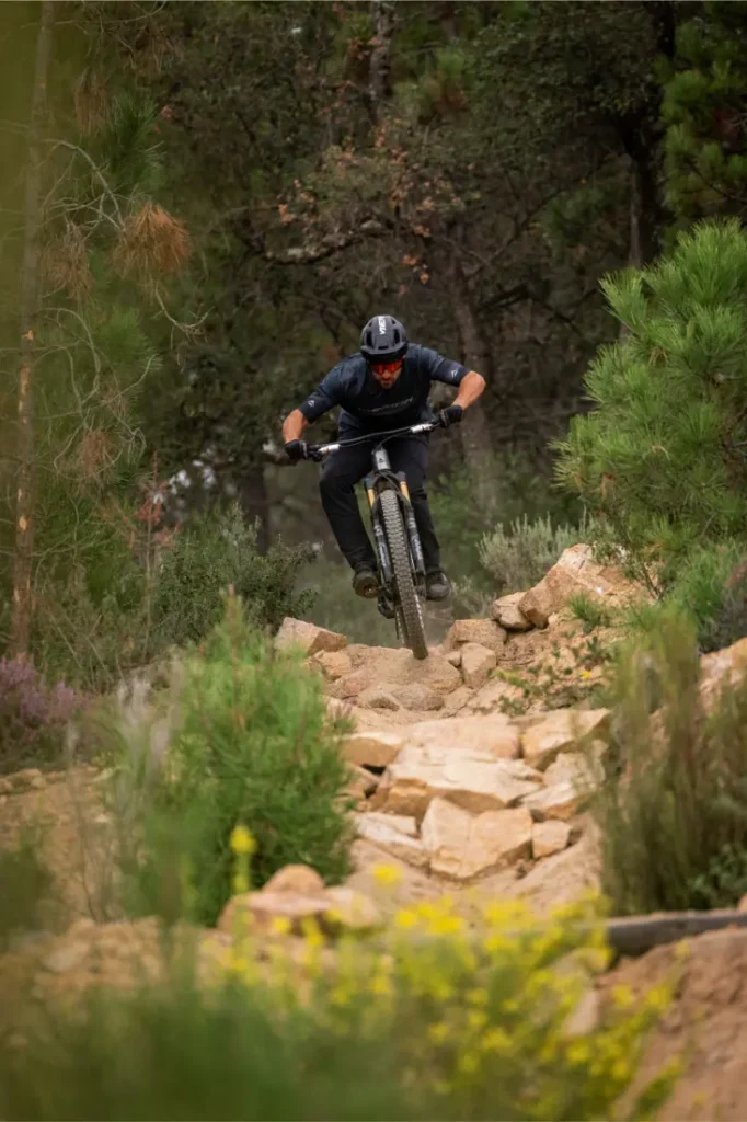 Una persona que lleva casco y equipo de protección va en bicicleta de montaña por un sendero de tierra rocoso, rodeado de árboles y arbustos.