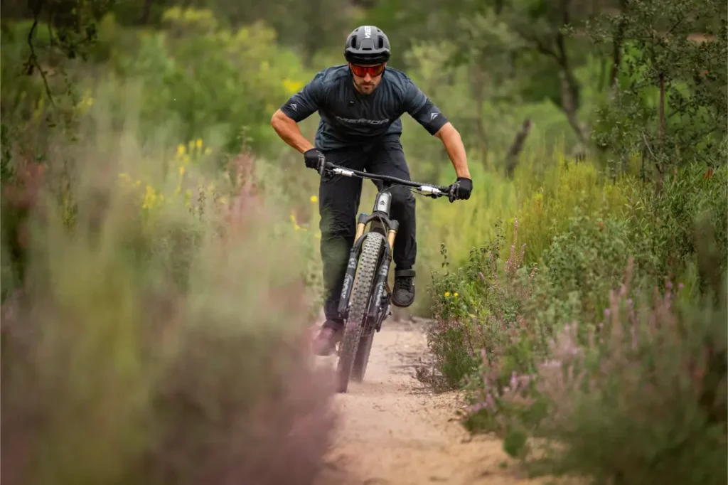 Una persona que lleva casco monta en bicicleta de montaña por un sendero de tierra rodeado de vegetación y flores silvestres.