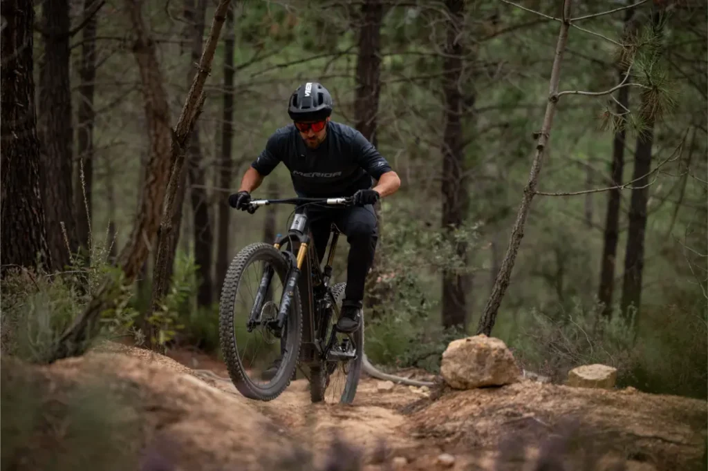 Una persona que lleva casco y gafas de sol sube en bicicleta de montaña por un sendero rocoso en una zona boscosa.
