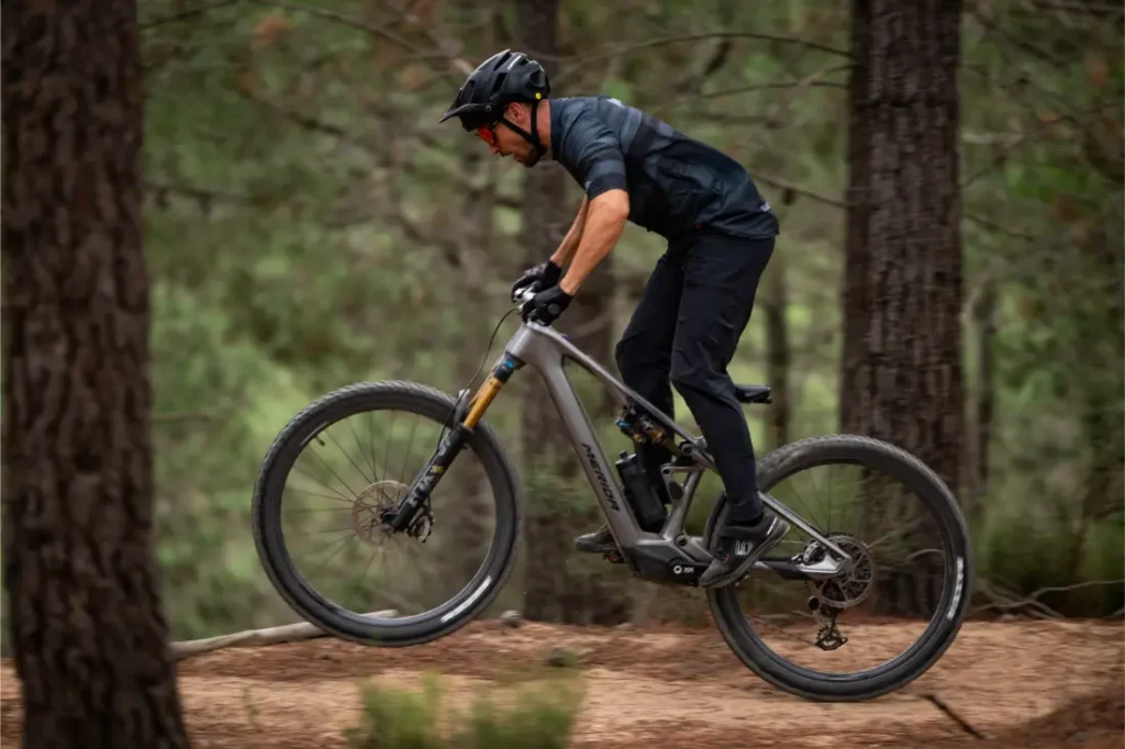Una persona que lleva casco y ropa oscura circula en bicicleta de montaña por un sendero de tierra en una zona boscosa, levantando la rueda delantera del suelo.