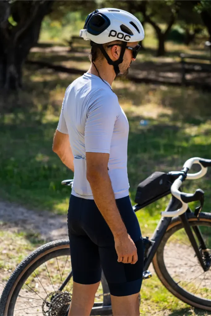 Una persona que lleva un casco polivalente y ropa de ciclismo posa junto a una bicicleta al aire libre en un día soleado, con árboles y césped al fondo, lista para una aventura por caminos de grava o por carretera.