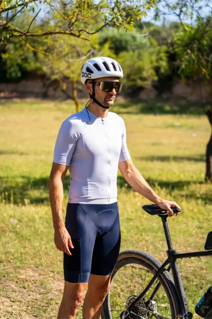 Un ciclista que lleva un casco polivalente blanco, gafas de sol y ropa de ciclismo está al aire libre junto a una bicicleta negra de gravel en un día soleado.