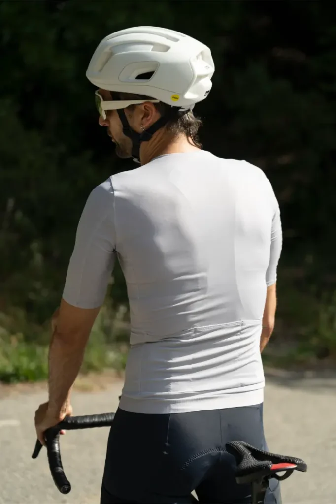 Un ciclista que lleva un casco blanco, gafas de sol y un maillot POC Cadence de manga corta en color gris claro circula por una carretera, visto desde atrás, disfrutando de la comodidad a la hora de pedalear que también ofrece el culote Cadence Cargo.