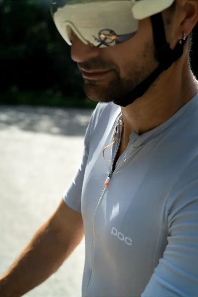 Un hombre que lleva un maillot blanco POC Cadence, un casco y unas gafas de sol reflectantes se encuentra al aire libre. La marca «POC» se ve claramente en su maillot, lo que destaca la comodidad a la hora de pedalear, ideal si se combina con el culote Cadence Cargo.