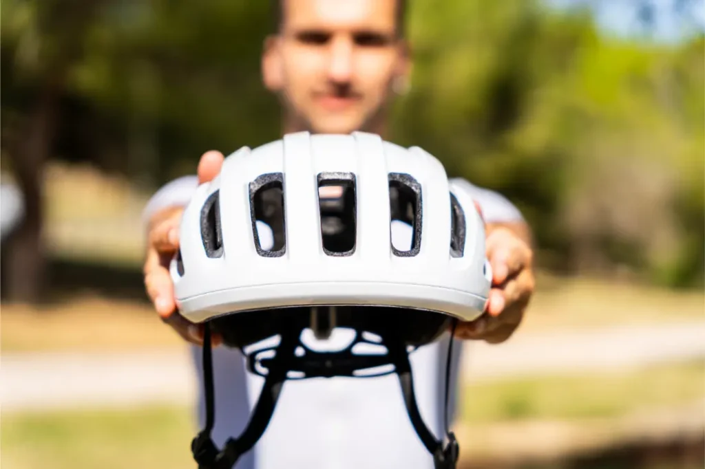 Una persona que sostiene un casco de bicicleta blanco «casco polivalente» hacia la cámara con los brazos extendidos, al aire libre con un fondo verde difuminado: protección perfecta tanto si vas por grava como por carretera.
