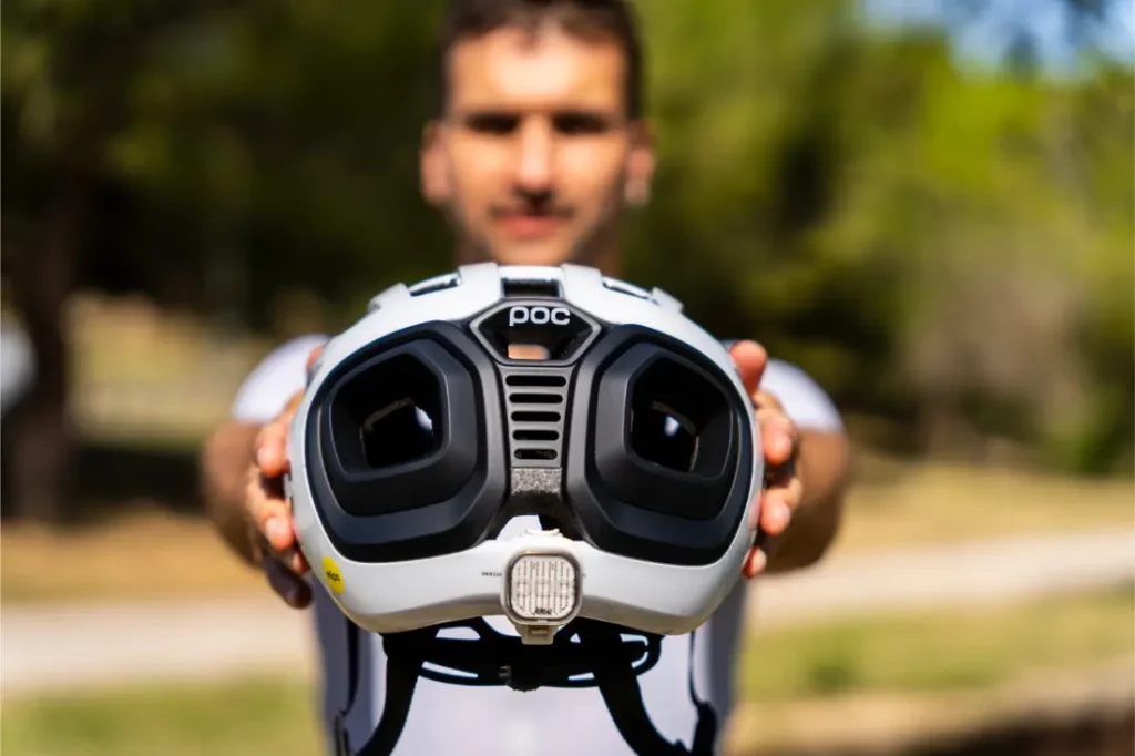 Una persona sostiene un casco polivalente blanco de la marca POC Amidal, con grandes aberturas de ventilación negras orientadas hacia la cámara, al aire libre y con vegetación al fondo, lo que lo hace ideal para el ciclismo de carretera.