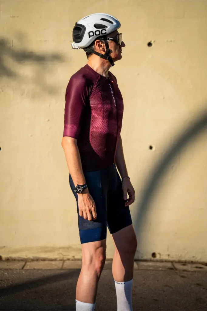 Un ciclista que lleva un casco blanco, gafas de sol, un maillot marrón rojizo Castelli Climber’s A/C y unos pantalones cortos tipo culotte azul marino A/C se encuentra de pie en una calle bañada por el sol, junto a una pared de color beige, preparado para dar el máximo rendimiento en condiciones de calor.