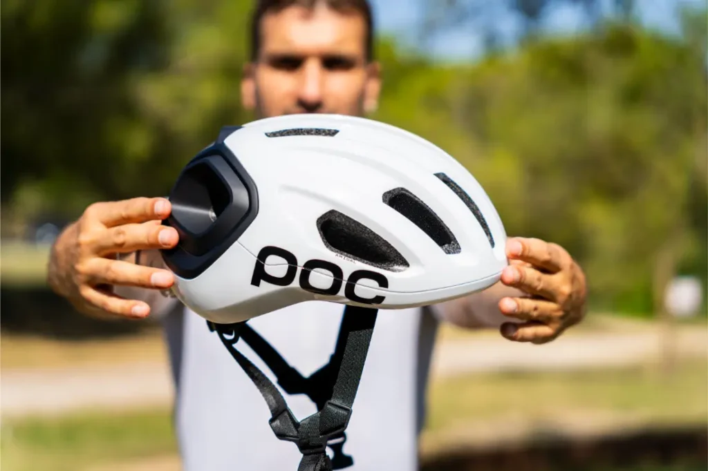 Una persona sostiene al aire libre un casco de ciclismo polivalente POC blanco con rejillas de ventilación negras y una correa para la barbilla, con árboles y césped al fondo; perfecto tanto para salidas por carretera como por grava.