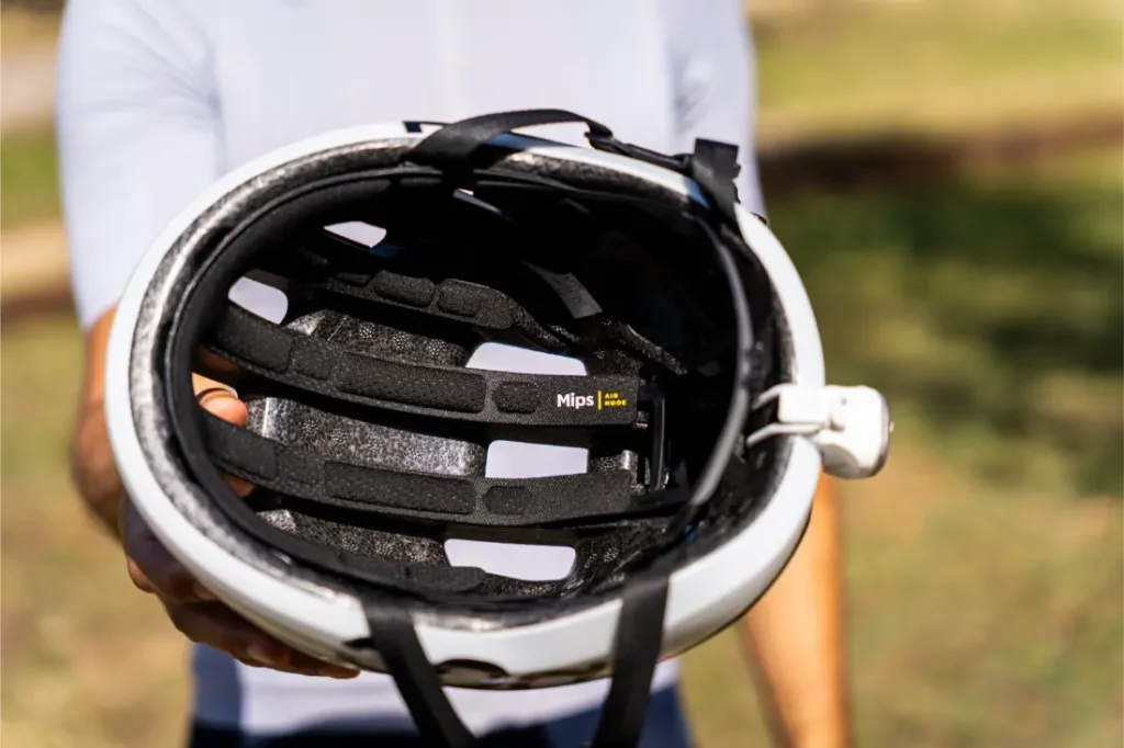 Una persona sostiene ante la cámara un casco de bicicleta blanco «Casco Polivalente», mostrando el acolchado interior y una etiqueta de seguridad amarilla con la inscripción «Mips» visible en el interior. El fondo es una zona al aire libre con césped, perfecta para aventuras en gravel o carretera.