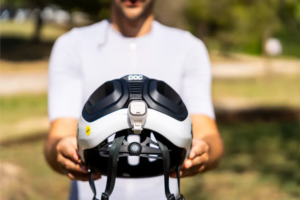 Una persona con camisa blanca sostiene un casco de bicicleta polivalente POC Amidal en blanco y negro, con una luz trasera incorporada, al aire libre en un día soleado: ideal tanto para aventuras por carretera como por grava.