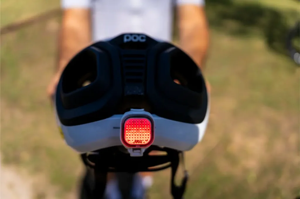 Primer plano de un casco de bicicleta polivalente con una luz trasera de seguridad roja acoplada, sostenido por una persona que se encuentra al aire libre en un día soleado; ideal tanto para aventuras en carretera como en grava.