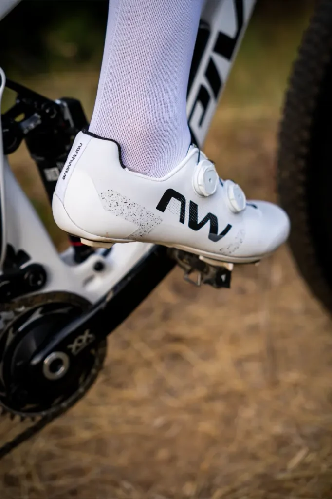 Primer plano del pie de un ciclista que lleva unas zapatillas blancas de ciclismo Northwave Extreme X en el pedal de una bicicleta, con parte de la bicicleta y hierba seca al fondo, lo que resalta la versatilidad y el diseño de alta gama del calzado.