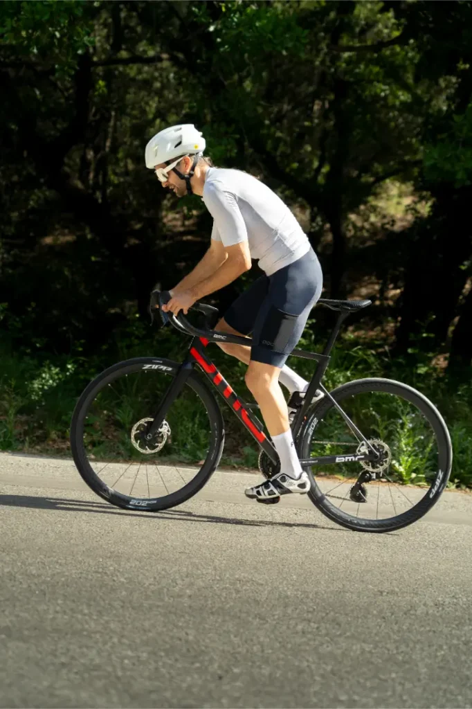 Una persona que lleva un casco blanco y ropa de ciclismo —con el maillot POC Cadence y el culote Cadence Cargo, para mayor comodidad al pedalear— circula en una bicicleta de carretera negra y roja por una carretera asfaltada, con árboles al fondo.