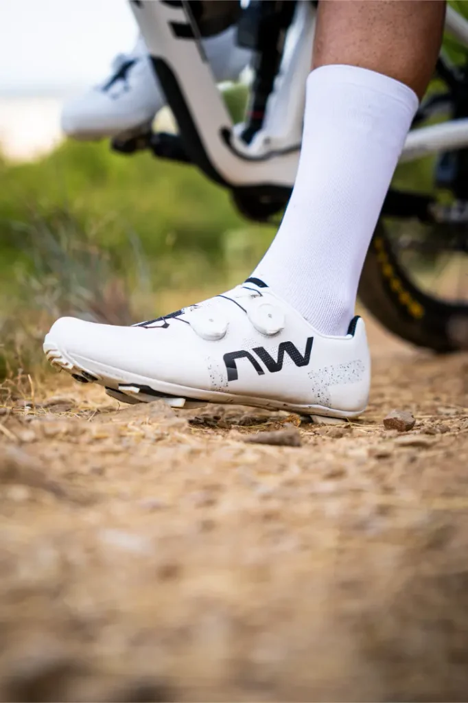 Una persona que lleva unas zapatillas de ciclismo blancas Northwave Extreme X, conocidas por su rendimiento de gama alta y su excepcional versatilidad, junto con unos calcetines blancos altos, está de pie en un camino de tierra junto a una bicicleta.
