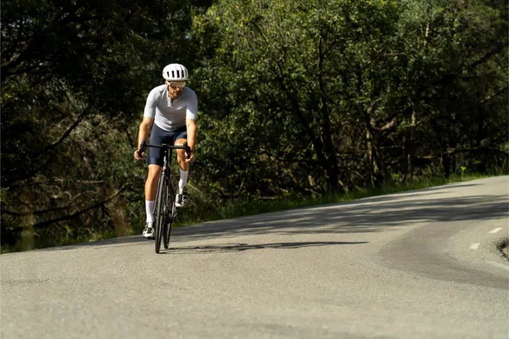 Un ciclista que lleva un casco blanco y ropa blanca —entre otras cosas, un maillot POC Cadence y un culote Cadence Cargo para mayor comodidad al pedalear— sube en bicicleta de carretera por una carretera sinuosa y bañada por el sol, bordeada de árboles.