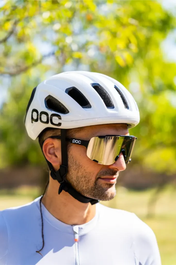 Una persona que lleva un casco de ciclismo polivalente blanco de la marca POC, gafas de sol y un maillot de ciclismo de color claro está de pie al aire libre, con árboles al fondo, lista para una aventura por carretera o por rutas de grava.
