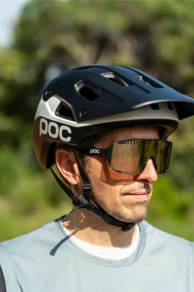 Una persona que lleva un casco de ciclista POC Cularis Pure en blanco y negro y unas gafas de sol con viseras grandes se encuentra al aire libre, disfrutando del sol con vegetación al fondo.