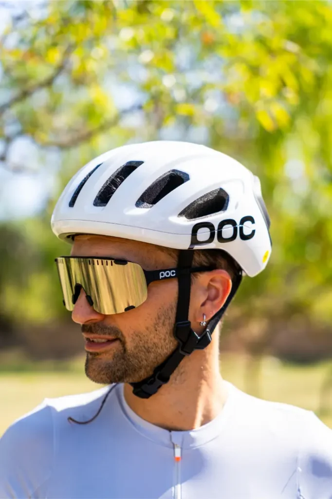 Una persona que lleva un casco de bicicleta blanco «casco polivalente» y unas grandes gafas de sol de espejo está de pie al aire libre, mirando hacia un lado, lista para una aventura por carretera o por rutas de grava.