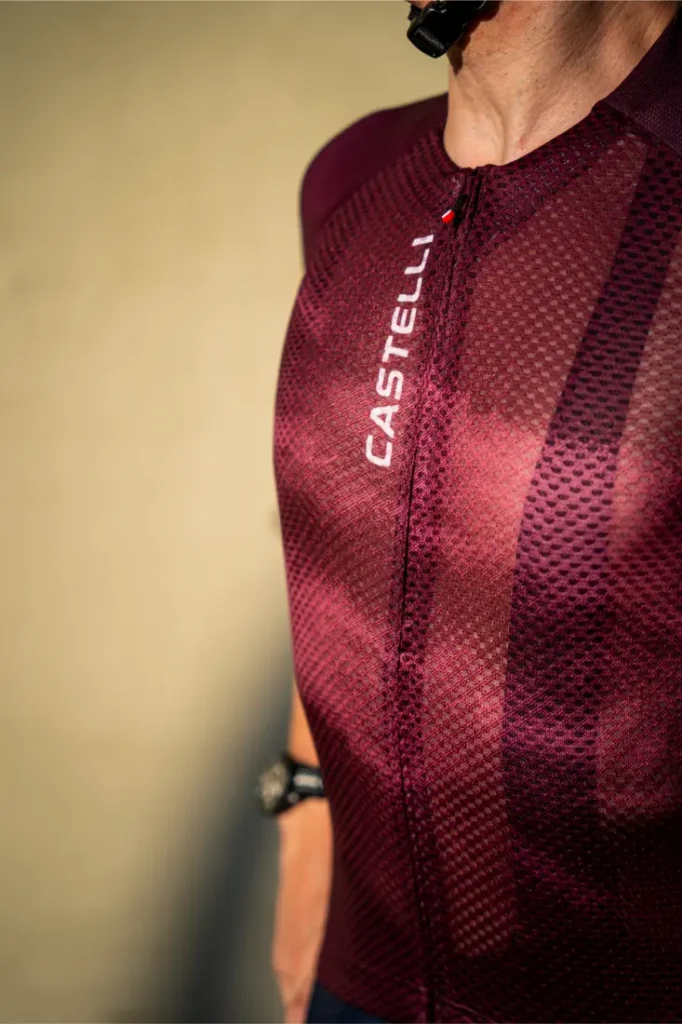 Persona que lleva un maillot de ciclismo Castelli de color granate y unos culotes Climber’s A/C, mostrada desde los hombros hasta la cintura, de pie sobre un fondo liso de color beige para un máximo rendimiento térmico.