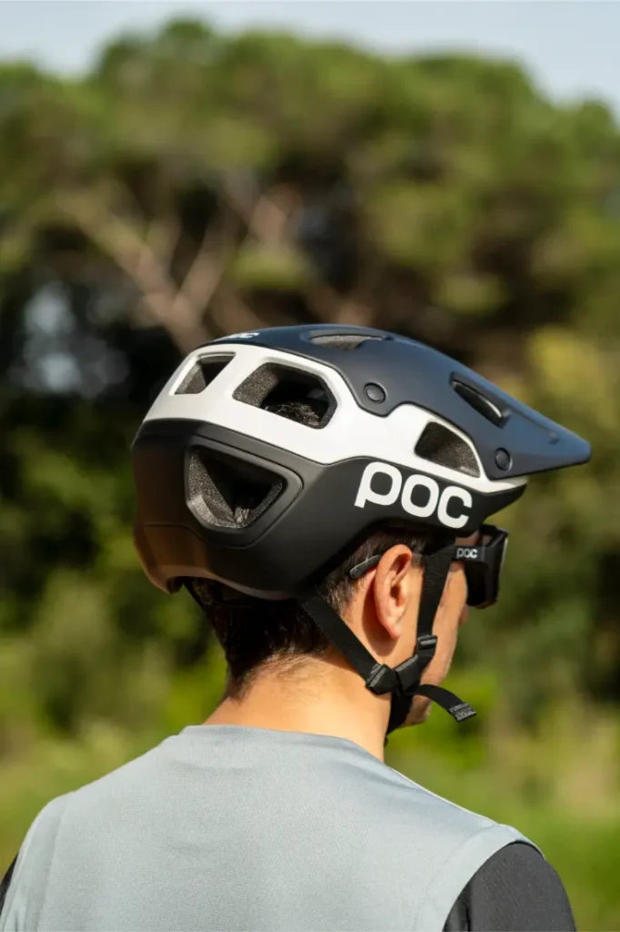 Se ve desde atrás a una persona que lleva un casco de bicicleta POC Cularis Pure, en blanco y negro, con viseras para protegerse del sol, de pie al aire libre con árboles al fondo.