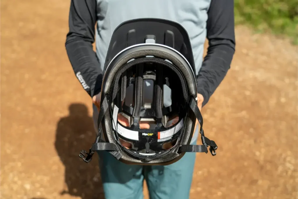 Una persona vestida con ropa de montaña sostiene frente a la cámara un casco de bicicleta negro POC Cularis Pure, mostrando claramente el acolchado interior, las correas y los detalles de la visera.