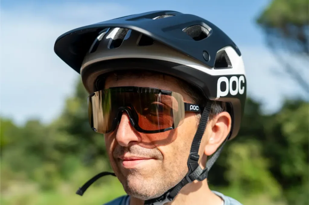 Una persona que lleva un casco de ciclista negro POC Cularis Pure y unas grandes gafas de sol de ciclista con cristales marrones se encuentra al aire libre, disfrutando del sol con una exuberante vegetación al fondo. Las elegantes viseras del casco aportan una protección adicional y un toque de estilo a su recorrido.