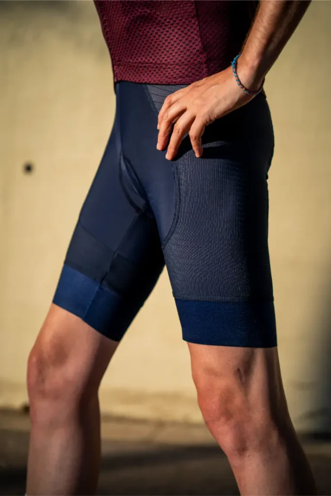 Una persona que lleva unos culotes de ciclismo Castelli Climber’s A/C de color azul marino y una camiseta de malla granate posa con una mano en la cadera ante un fondo liso, preparada para dar el máximo rendimiento en condiciones de calor.