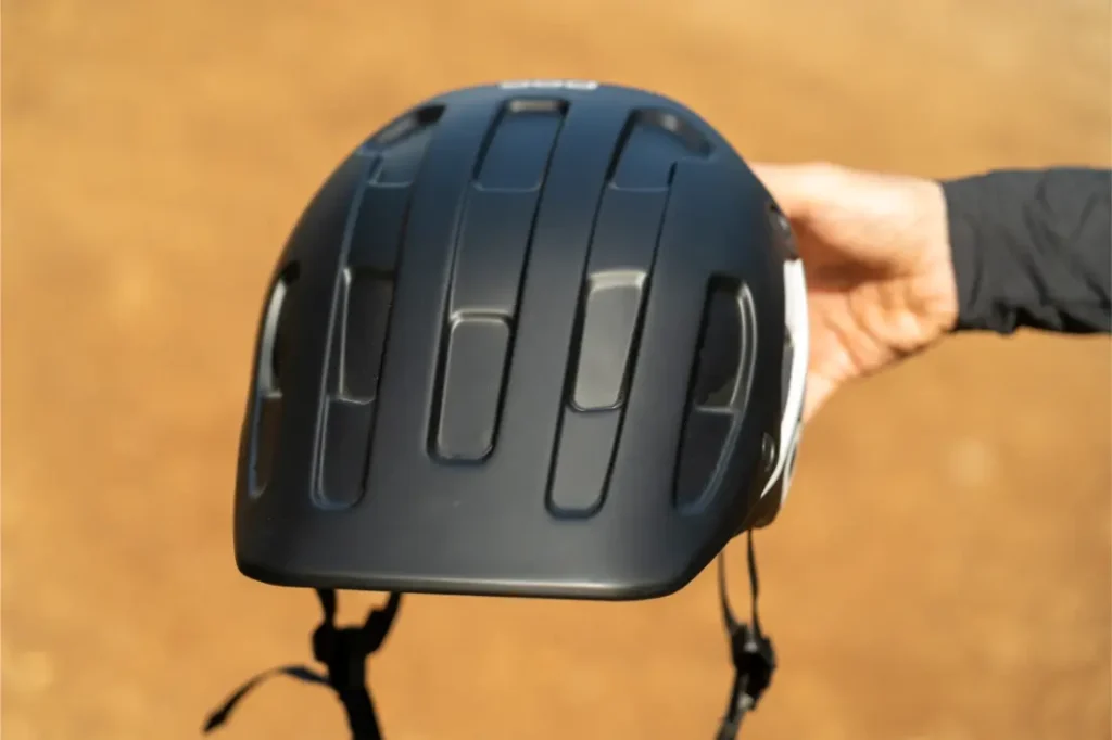 Una persona sostiene un casco de bicicleta POC Cularis Pure negro, con múltiples rejillas de ventilación y una correa ajustable, que se muestra al aire libre sobre un fondo marrón difuminado. El casco cuenta con viseras Sol para una mayor protección y comodidad durante los recorridos bajo el sol.