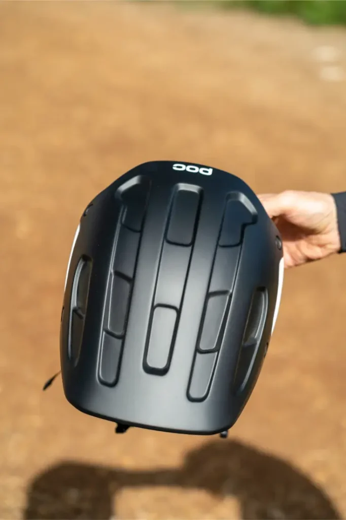 Una mano sostiene un casco de ciclismo POC Cularis Pure negro con acabado mate y ranuras de ventilación verticales, diseñado para adaptarse al clima, al aire libre en un camino de tierra.