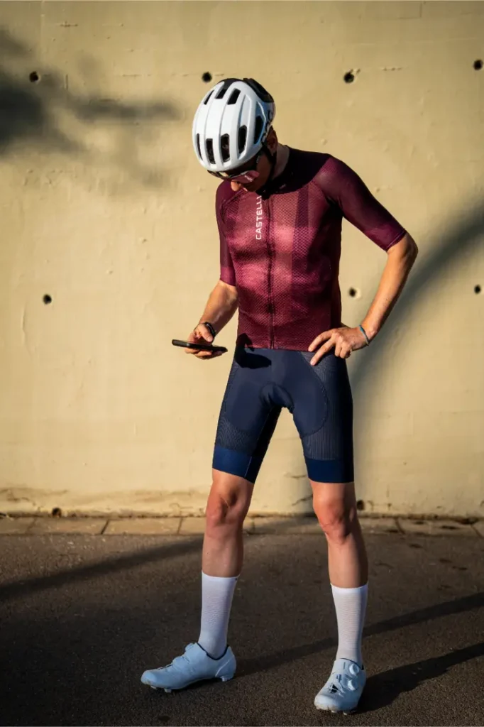 Un ciclista con casco y equipación de ciclismo, vestido con un maillot Castelli Climber y unos culotes A/C para un rendimiento térmico óptimo, está de pie en la acera mirando el móvil que tiene en la mano, con sombras y una pared de color beige al fondo.