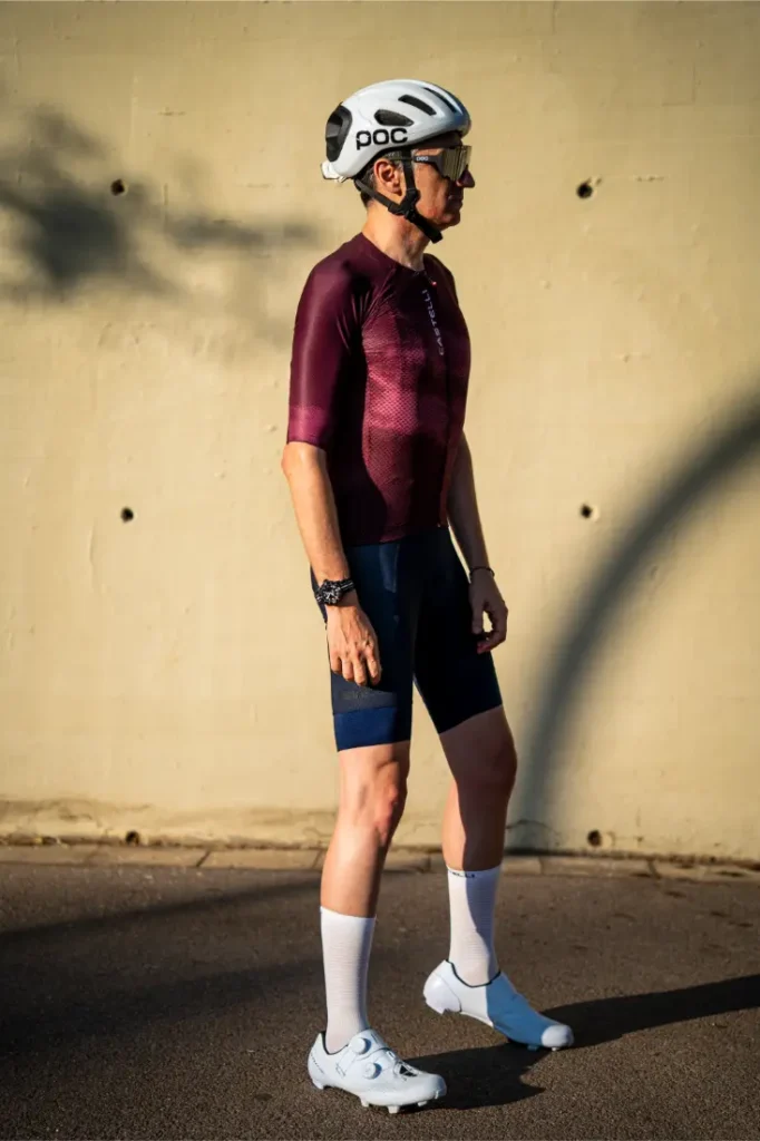 Un hombre que lleva un maillot de ciclismo marrón rojizo de la marca Castelli Climber’s, unos pantalones cortos tipo culotte azul marino de la marca A/C, un casco blanco, calcetines blancos y zapatillas de ciclismo está de pie sobre una superficie pavimentada junto a una pared de color beige, preparado para dar el máximo rendimiento bajo el calor.