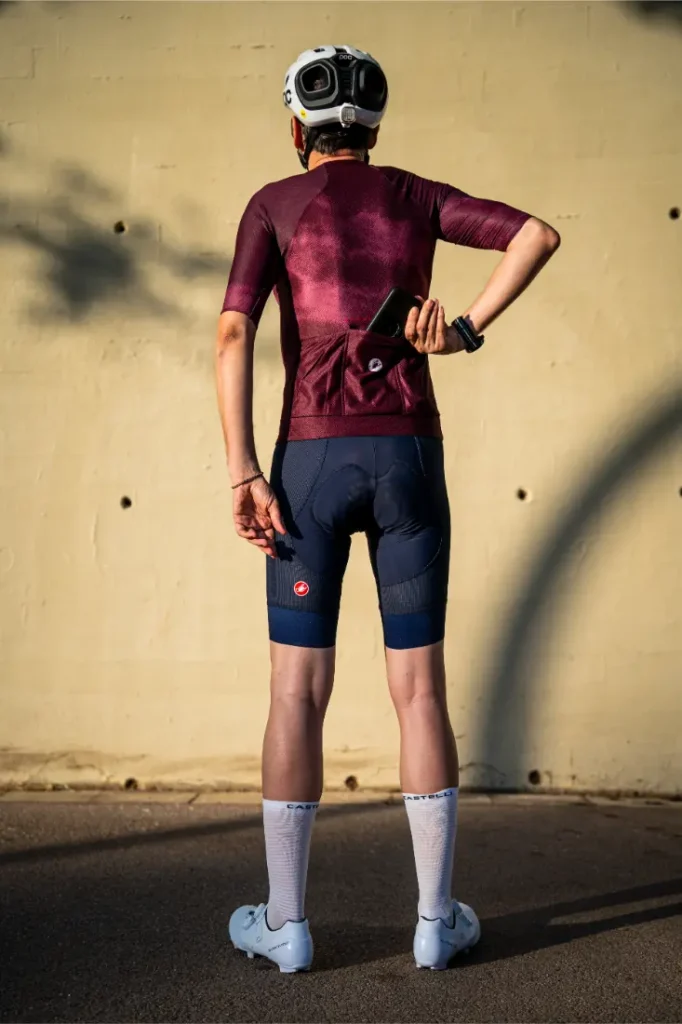 Un ciclista con maillot granate, pantalones cortos tipo culotte A/C azul marino, calcetines blancos y casco está de pie frente a una pared, guardándose el móvil en el bolsillo trasero, listo para rendir al máximo gracias al conjunto Castelli Climber’s A/C.