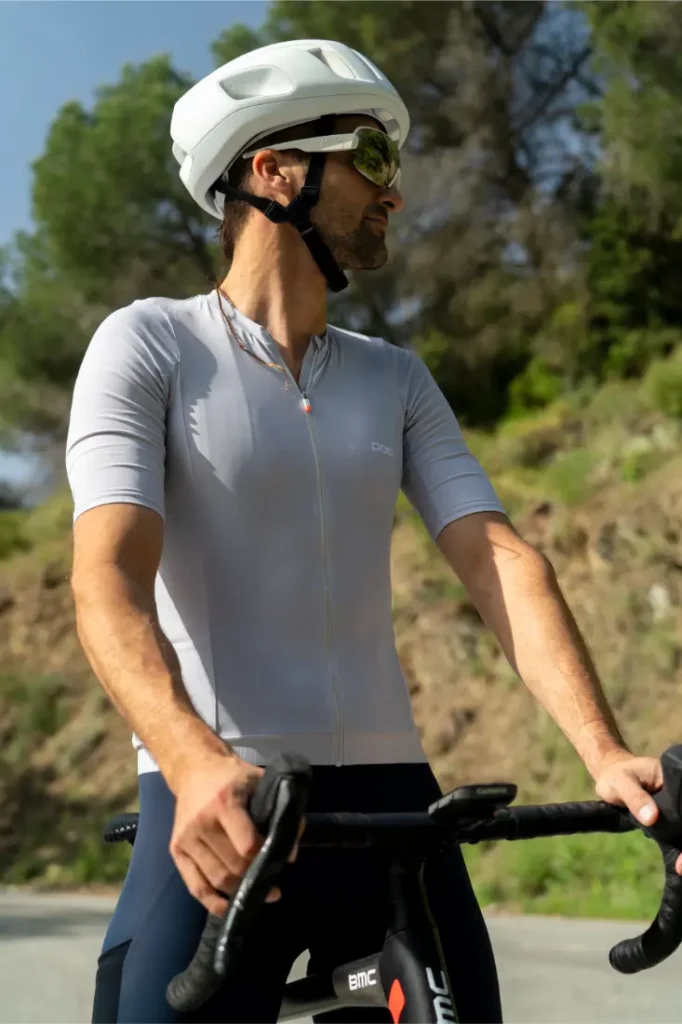 Una persona que lleva casco, gafas de sol y un maillot de ciclismo POC Cadence de color gris claro va en bicicleta al aire libre en un día soleado, con árboles al fondo, disfrutando de la comodidad para pedalear que le proporciona su culotte Cadence Cargo.