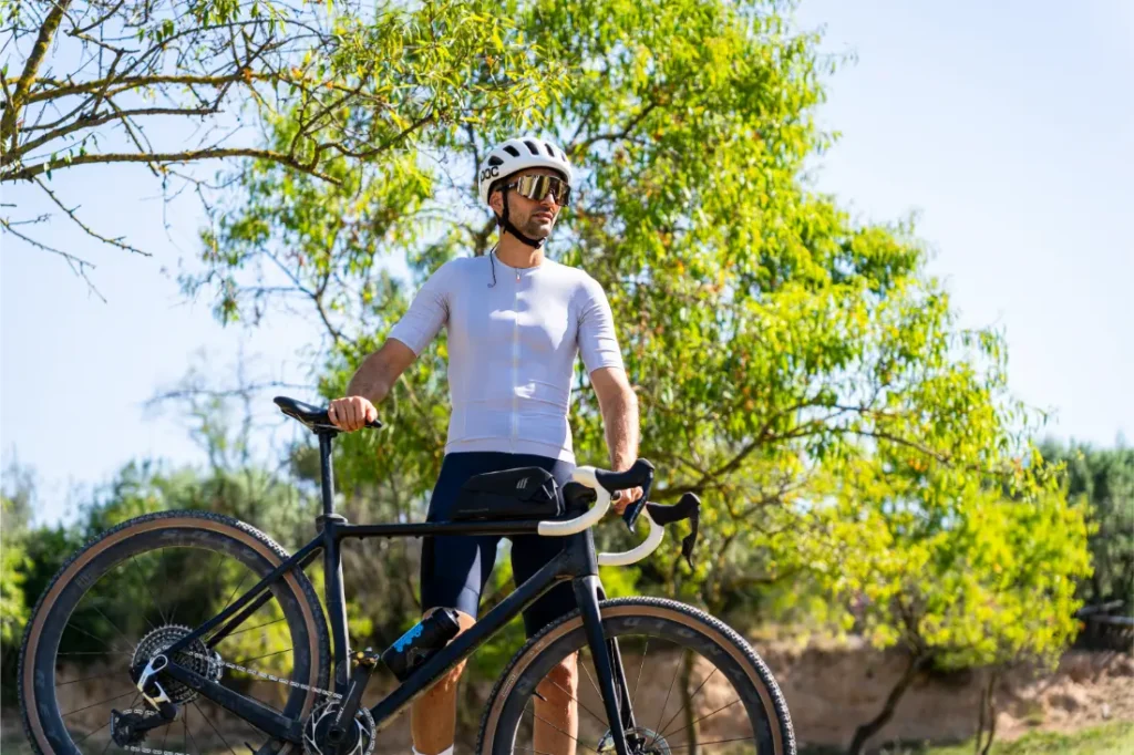 Un ciclista que lleva un casco polivalente blanco y ropa de ciclismo está al aire libre junto a una bicicleta Gravel negra, rodeado de árboles verdes.